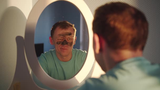 Молодой Человек Снимает Лица Черную Косметическую Маску Глядя Себя Зеркало — стоковое видео