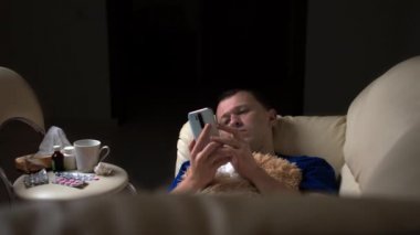 Genç adam hasta, kanepede tek başına yatıyor ve telefonu kullanıyor.