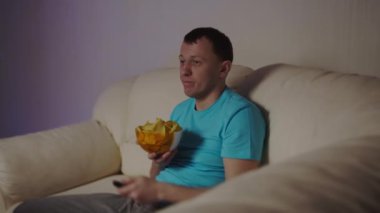 Adamın biri TV 'nin yanındaki koltukta oturup cips yiyor.