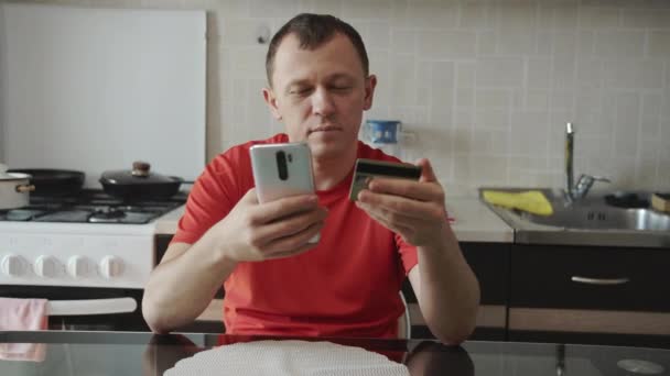 Un hombre sentado en la cocina paga por una compra en línea a través de un teléfono móvil — Vídeo de stock