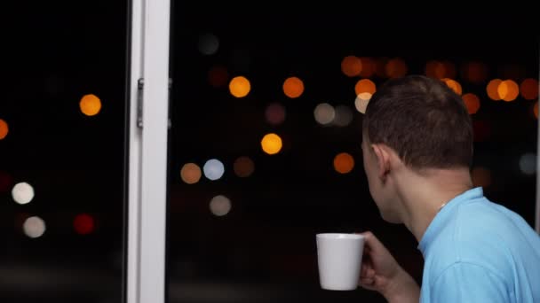 Genç Adam Sıcak Bir Içecek Içer Pencerenin Dışındaki Işıklara Bakar — Stok video