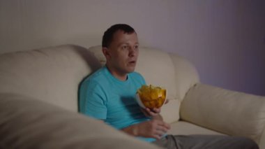 Adam TV 'de gördüğü yayına şok oldu, elinde bir kase cipsle kanepede oturuyor.