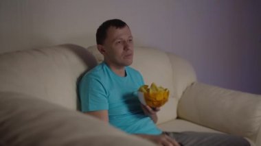 Akşamları kanepede oturup cips yerken komik bir program izleyen bir adam.