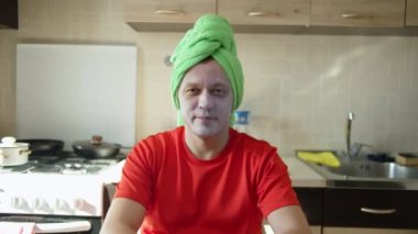 Yüzünde kozmetik bir maske ve kafasında bir havluyla oturan bir adam.