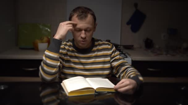 Joven con expresión lee un libro mientras está sentado en una mesa por la noche en la cocina — Vídeo de stock