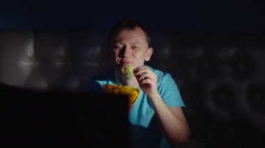 Gecenin geç saatlerinde elinde bir kase cipsle neşeli bir adam TV izliyor, kamera hareket ediyor.