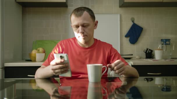 男は台所に座ってお茶を飲みながら携帯電話でメッセージを読み取ります — ストック動画