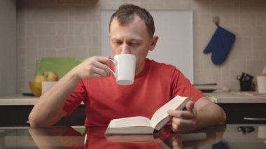 Adam güneşli bir günde mutfakta otururken çay içiyor ve kitap okuyor.