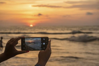 Endonezya, Bali 'deki Kuta plajına yakın bir yerde cep telefonuyla dramatik bir gün batımı fotoğrafı çeken kadın gezgin.