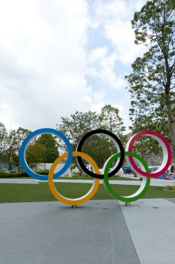 SHINJUKU CITY, TOKYO, JAPAN - SEPTEMBER 30, 2019: Front view of Olympic Rings. Background with New National Stadium (Tokyo) and the Cauldron (The XVIII Olympic Games Nagano 1998). Vertical shot. clipart