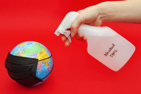 Hand spraying 70% alcohol on terrestrial globe model with black surgical mask isolated on red background. Concept: Quarantined earth. Protection against Coronavirus (COVID-19) SARS-CoV-2. Horizontal shot.