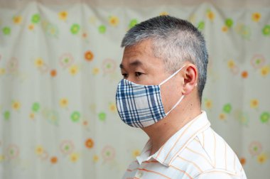 Coronavirus (COVID-19) ve diğer bulaşıcı hastalıklara karşı korunmak için ev yapımı kumaş yüz maskesi takan orta yaşlı bir Asyalının profil yan görüntüsü. Çiçekli perde arkası. Boşluğu kopyala.