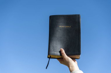 Raised hand holding the Holy Bible. Background with blue sky on a beautiful winter morning. Copy space. Close-up. Horizontal shot. clipart