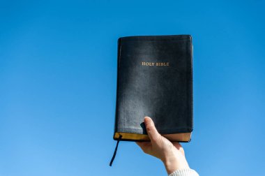 Raised hand holding the Holy Bible. Background with blue sky on a beautiful winter morning. Copy space. Close-up. Horizontal shot. clipart