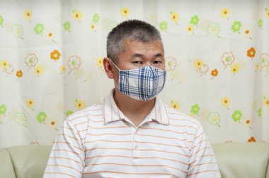 Coronavirus (COVID-19) ve diğer bulaşıcı hastalıklara karşı korunmak için maske takan orta yaşlı Asyalı bir adamın kanepeye oturması ön planda. Çiçek perde arkaplanı.