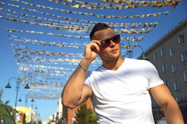 Güneş gözlüklü ve beyaz tişörtlü bir adam başka tarafa bakıyor. Mavi gökyüzündeki çok renkli bayrakların arka planında