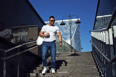 Beyaz tişört, kot pantolon ve beyaz spor ayakkabı giyen bir adam merdivenlerden sırt çantasıyla iniyor.