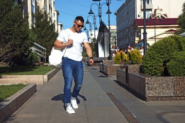 Sokakta elinde mavi kot pantolon, beyaz t-shirt ve güneş gözlüğü olan genç bir adam yürüyor.