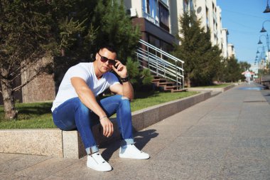 Sokakta oturan genç yakışıklı adam. Beyaz tişört, kot pantolon, moda ayakkabısı, güneş gözlüğü, hippi tarzı adam.