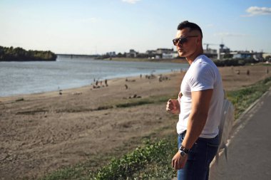 Güneş gözlüklü, beyaz tişörtlü ve mavi kotlu bir adam sırtında sırt çantasıyla nehrin kenarında duruyor. Yan görünüm