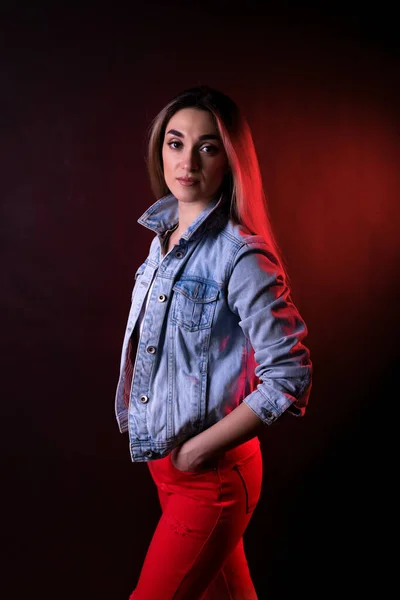 A young girl in a denim jacket and red trousers on a black background with red lighting