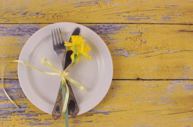 Yer belirleme konsepti. Ahır tahtalarında çatal bıçak takımı olan bahar tatili masası. Beyaz tabakta sarı narsisli bant ile bağlanmış çatal bıçak yemek. Eski sarı ahşap masa, fotokopi alanı ile üst manzara.