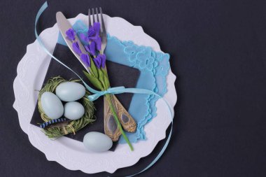 Siyah arka planda çatal bıçak seti olan güzel bir Paskalya tatili masası. Mor çiçek, yumurta, Tiffany renk peçete ve beyaz tabakta fiyonklu mutfak aletleri. Kopyalama alanı olan tek renkli daire.