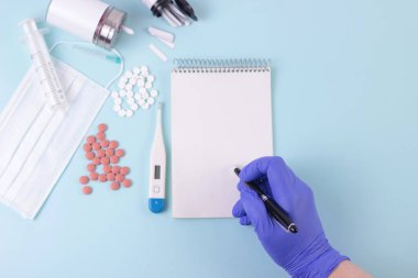 İlaç, eczane, sağlık konsepti. Doktorlar mavi tıbbi eldiven verip reçete yazıyorlar. İçinde haplar, termometr, ilaç maskesi, şırınga olan mavi masa. Boş not defterini taklit et. Üst görünüm.