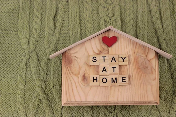 The inscription stay home on little wooden house lying on a knitting green plaid . The concept of staying at home to avoid spreading the coronavirus. Top flat lay with copy space. — Stock Photo, Image
