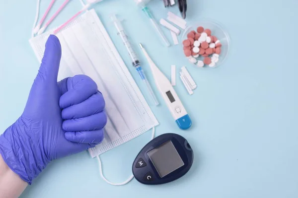 Medicine, control of blood sugar levels.Thumb up approval symbol. Hand in blue latex glove showing OK sign: good test results.Medical background: pills, mask, thermometr, test strips and glucometer.