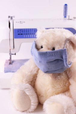 Beyaz arka planda dikiş makinesinde ev yapımı mavi maske takan yumuşak bir oyuncak ayı. Kapatın. Karantina ve viral enfeksiyonun önlenmesi kavramı.