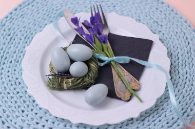 Güzel Paskalya bayramı sofrası, çatal bıçak takımı ve beyaz tabakta mor çiçekler. Yuvarlak örülmüş turkuaz renkli peçete. Yemek masası. Güzel bir yemek restoranı. Seçmeli odak noktası.