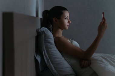 Genç bir kadın gece geç saatlerde telefonunu bağımlılık yapıcı bir şekilde kullanıyor.