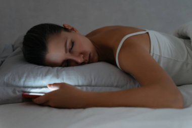 Geç saatte elinde akıllı telefonuyla yatakta uyuyan genç bir kadın. 