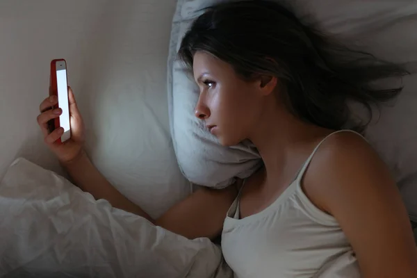 Jong wijfje liggend in bed en kijkend naar haar smartphone op bijna — Stockfoto