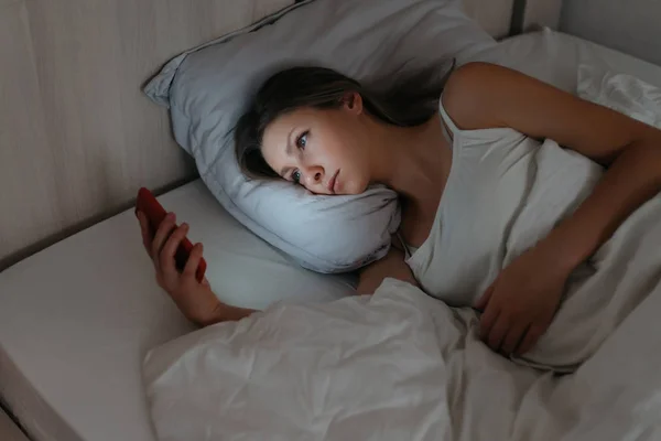 Fiatal nő feküdt az ágyban, és nézi az okostelefon éjjel. — Stock Fotó