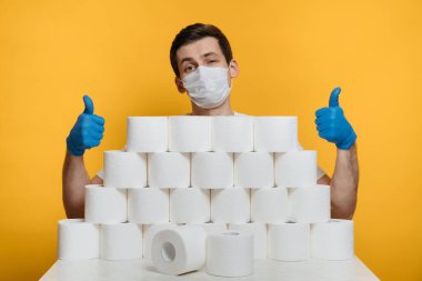 Kendine güveni tam, koruyucu maskeli ve tuvalet kağıdı yığınının arkasında duran tıbbi eldivenli mutlu genç adam. Evde kaldığın için koronavirüsten kendini koru. Covid 19