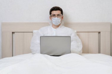 Biyolojik tehlike giysileri içindeki korkmuş genç adam Coronavirus karantinası sırasında dizüstü bilgisayarıyla evden uzaktan çalışıyor. Covid-19 enfeksiyonlu hasta kendini izole ediyor. Virüs salgını. Sosyal uzaklık