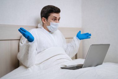Biyolojik tehlike giysisi giymiş genç beyaz bir adam dizüstü bilgisayar kullanıyor ve koronavirüs enfeksiyonu yayıldığı haberiyle şok oluyor. Covid-19 karantinası yüzünden işini kaybetti. Evde kal ve sosyal mesafe kavramı
