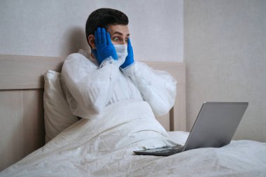 Biyolojik tehlike giysileri içindeki genç beyaz adam dizüstü bilgisayar kullanıyor ve kendini izole bir şekilde evde kalıyor COVID-19 salgını hakkındaki bilgilerle şoke oluyor. Coronavirüs