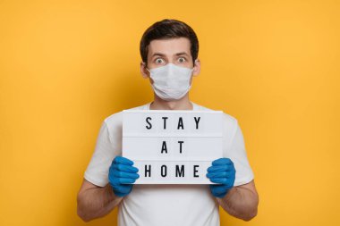 Tıbbi maskeli ve eldivenli korkmuş bir adam. Evde mesajda kalın yazıyor. Covid-19 veya Coronavirus konsepti. Sarı arka planda izole edilmiş. Coronavirus önleme için sosyal medya kampanyası