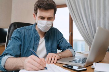 Coronavirus ya da COVID 19 salgını yüzünden tıbbi yüz maskeli genç bir adam dizüstü bilgisayarla uzaktan çalışıyor. Uzaktan eğitim ve serbest çalışma. Evde kal konsepti. Karantina sırasında kaybedilen iş