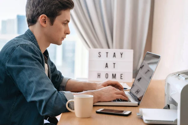 Young caucasian man working remotely with laptop and smartphone in his room. Freelance work and electronic learning. Lost job due to COVID-19 pandemic and quarantine. Board with Stay home text