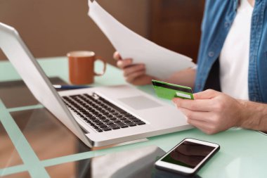 Kafkasyalı genç işadamı, ev işlerinde bilgisayar, akıllı telefon ve kredi kartı kullanıyor. Çevrimiçi alışveriş ve e-bankacılık konsepti. İnternete para yolladım, rezervasyon