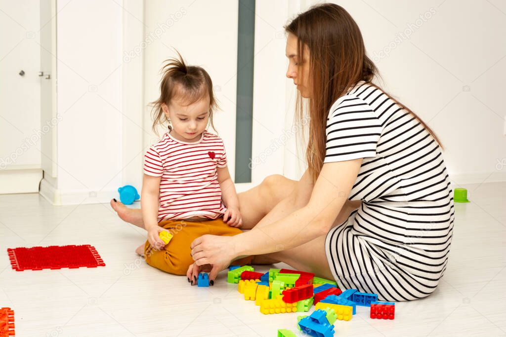 Little child girl and her mother sitting on the floor and playing together with colorful constructor block