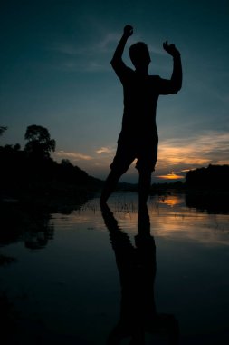 Güzel bir gün batımının önünde poz veren bir adamın silüeti güzel manzaralı nehrin suyunu yansıtıyor.