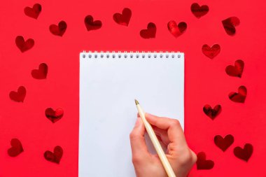 Kırmızı arka planda elinde kalem olan, beyaz, boş bir not defteri ve kırmızı kalpli bir konfeti.