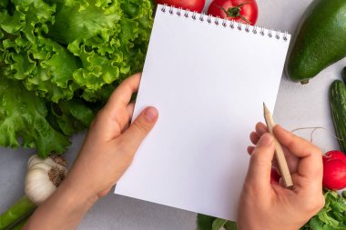 Sebze arka planında kağıt not defteri ve kalem tutan kadın elleri. Alkali ve vejetaryen diyeti