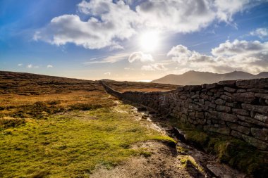 Slieve Donard dağının kıyısındaki yas duvarı Mavi gökyüzü, beyaz bulutlar ve güneş ışınları.