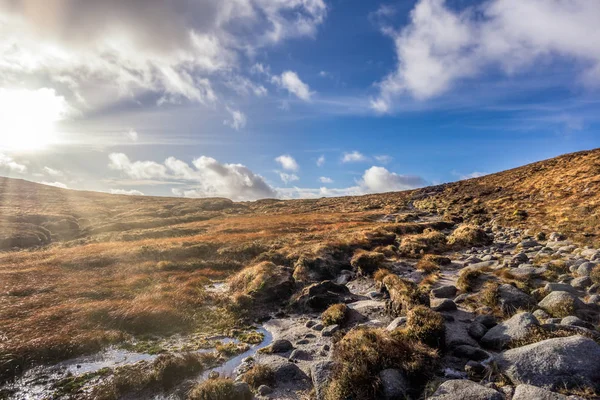Campo húmedo y rocoso en la orilla de la montaña Slieve Donard con cielo azul, nubes blancas y rayos de sol —  Fotos de Stock